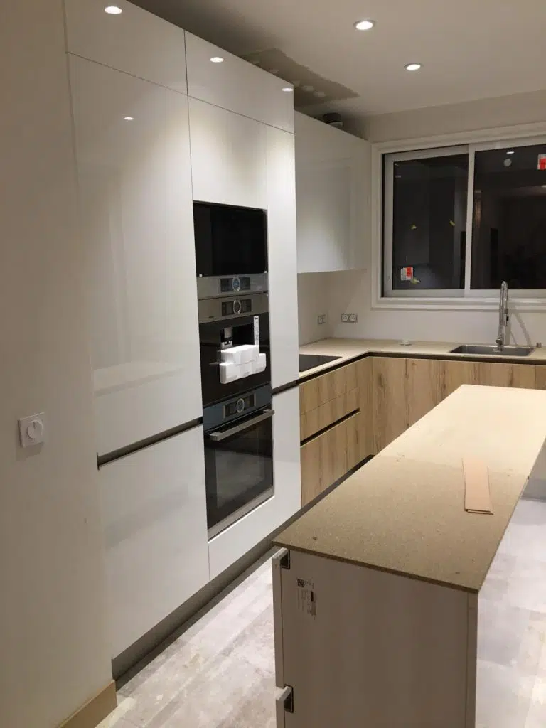 Découvrez notre dernière réalisation de cuisine, alliant fonctionnalité et esthétique moderne. Cette cuisine met en valeur des meubles bas effet bois clair, offrant une ambiance chaleureuse et naturelle. Les façades sans poignée avec gorge créent des lignes épurées et un design contemporain. Les meubles hauts laqués blancs apportent une touche de luminosité et de sophistication, contrastant élégamment avec le bois des éléments bas. L'îlot central, encore en cours de finition avec son plan de travail provisoire, promet un espace convivial et pratique pour la préparation et les repas. Chaque détail a été pensé pour créer une cuisine à la fois belle, pratique et durable, répondant aux exigences des modes de vie contemporains.