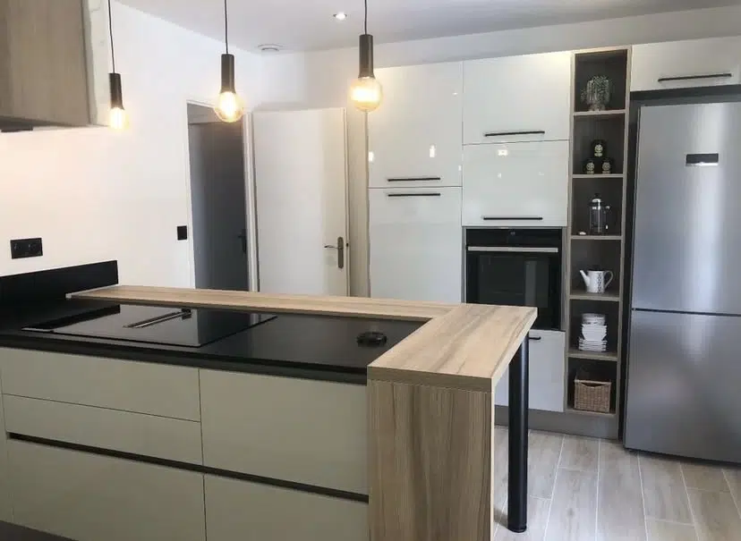 Découvrez cette réalisation qui allie à merveille esthétisme contemporain et fonctionnalité optimisée. Au cœur de cette cuisine, le contraste est saisissant entre le plan de travail noir et l'élégance des façades claires, créant une atmosphère chic et intemporelle. L'agencement en L, complété par un îlot central multifonctionnel, offre un espace de travail généreux et convivial. Cet îlot, habillé d'un comptoir en chêne clair de chez finitop, devient un lieu privilégié pour la préparation des repas comme pour les moments de partage informels. Les luminaires suspendus au-dessus de l'îlot apportent une touche chaleureuse et design. Nous avons intégré des solutions de rangement intelligentes avec des meubles hauts et bas épurés, ainsi qu'une colonne ouverte astucieuse pour exposer vos plus beaux objets. L'électroménager encastré s'intègre parfaitement, garantissant une ligne épurée et moderne. Cette cuisine est une invitation à la convivialité et à la praticité, pensée pour répondre aux exigences de la vie quotidienne.
