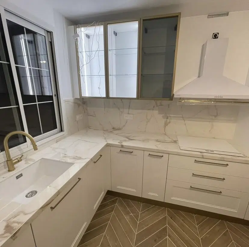 Découvrez l'élégance intemporelle et la luminosité éclatante de cette cuisine, une nouvelle réalisation qui capture l'essence du design classique revisité. Les façades meg, d'un blanc pur, offrent une toile de fond lumineuse et épurée, mettant en valeur la beauté naturelle du marbre. Leurs lignes classiques, agrémentées de poignées fines et dorées, rappellent une élégance intemporelle, tandis que les rangements sont pensés pour une organisation optimale. Les meubles hauts vitrés, encadrés de doré, allègent l'espace et offrent une opportunité d'exposer de la vaisselle raffinée, ajoutant une dimension décorative et aérienne à l'ensemble.