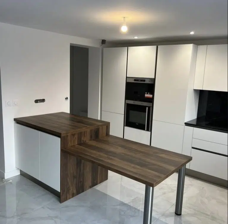 Découvrez l'une de nos récentes réalisations : une cuisine où le design contemporain rencontre une fonctionnalité intelligente. Cette création se distingue par l'alliance réussie du blanc épuré des façades Arredo3 et de la chaleur du bois foncé Fidelem. Cette combinaison intemporelle apporte une luminosité et une ambiance accueillante à l'espace. Au cœur de cette cuisine, l'îlot central se prolonge astucieusement en un coin repas convivial. Cette configuration optimisée offre à la fois une vaste surface de travail et un lieu de partage idéal. L'intégration parfaite de l'électroménager et des solutions de rangement discrètes témoigne de notre souci du détail et de notre engagement pour une ergonomie optimale. Chaque élément est pensé pour une utilisation fluide et un environnement ordonné.