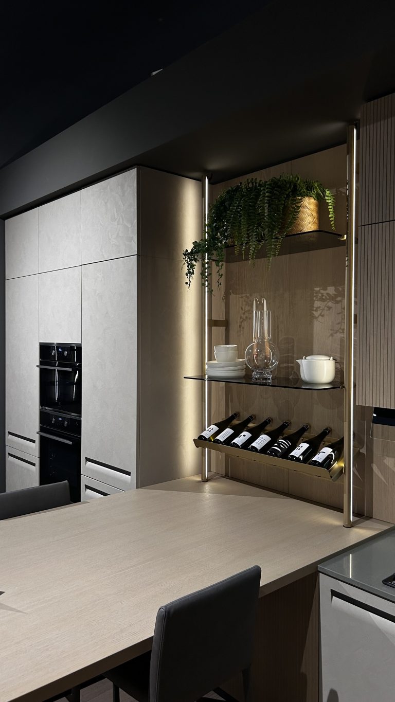 Découvrez une cuisine qui marie astucieusement design contemporain et solutions de rangement intelligentes. Les façades aux teintes claires et texturées s'accordent parfaitement avec les éclairages LED intégrés, créant une ambiance moderne et chaleureuse. Le meuble de rangement ouvert, doté d'un casier à bouteilles et d'étagères en verre, offre un espace d'exposition élégant pour vos objets et votre cave à vin. L'intégration harmonieuse des fours encastrés et le vaste plan de travail font de cette cuisine un espace aussi esthétique que pratique.
